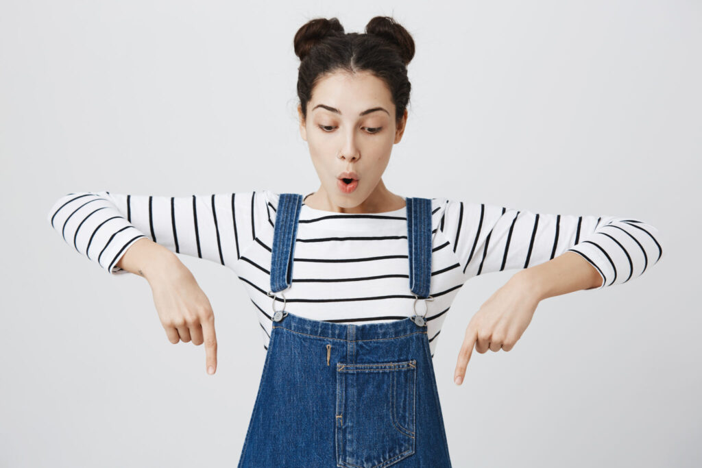 Excited surprised girl with hairbuns in denim overalls, pointing index fingers down, showing copy space for promotional content. Amazed funny female model advertising something